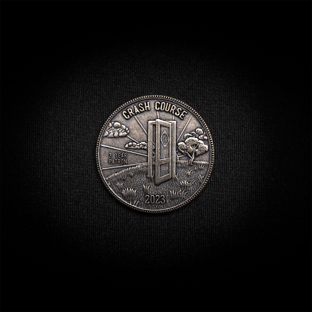 Image of the front side of a hand-engraved coin on a black background. The design on the coin is an image of a door frame with an open door on a grassy hill with sunshine rays beaming in the back. The coin is carved with the Crash Course title, the year 2023, and the phrase "3 year patron".